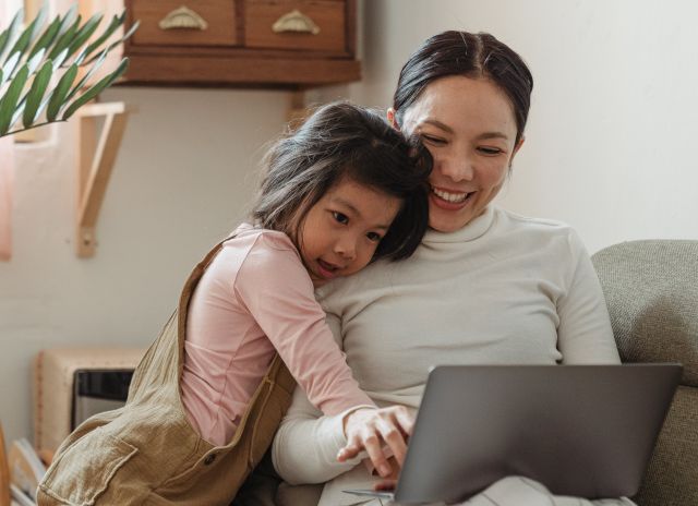 Cheerful smiling Asian woman browsing modern netbook while hugging with cute content daughter on comfy sofa in cozy living room